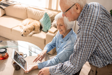 High angle  portrait of modern senior couple using digital tablet sitting at table at home with pet dog in background, copy space