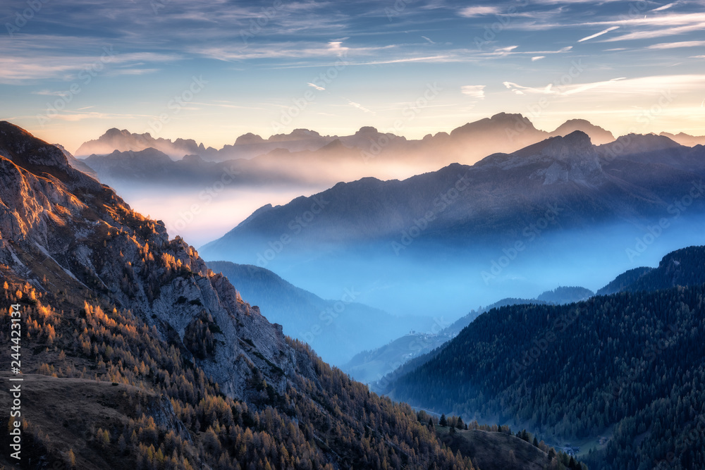 Fototapeta Góry w mgle przy pięknym zmierzchem w jesieni w dolomitach, Włochy. Krajobraz z alpejską górską doliną, niskie chmury, drzewa na wzgórzach, wioska we mgle, błękitne niebo z chmurami. Widok z lotu ptaka. Passo Giau