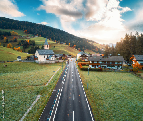 Aerial view of the road in beautiful village in mountain valley at sunrise. Dolomites, Italy. Top view of asphalt roadway, church, buildings, meadows with green grass, trees in autumn. Landscape