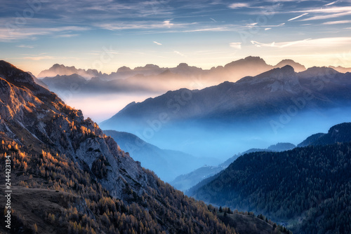 Mountains in fog at beautiful sunset in autumn in Dolomites, Italy. Landscape with alpine mountain valley, low clouds, trees on hills, village in fog, blue sky with clouds. Aerial view. Passo Giau