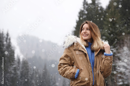 Young woman in warm clothes near forest, space for text. Winter vacation