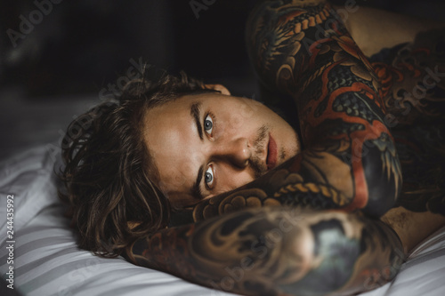 Young handsome tattooed man just woke up and laying in bed in the bedroom.
