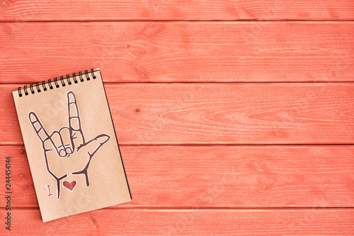 A notebook of Kraft paper with a painted symbol ASL (American Sign Language) ILY (I Love you) that lies on the textured wooden surface of the color 
