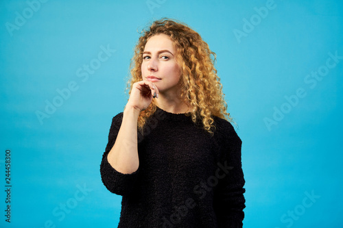 Portrait of thoughtful attractive redhead curly girl looking at camera dressed in black sweater on blue background.