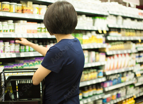 Woman Shopping at Grocery Market Pharmacy. Supermarket Shopper Doing Groceries. Female Holding Basket Trying to Decide which Products to Buy. Retail Healthcare Medicine, Vitamins, and Supplements.