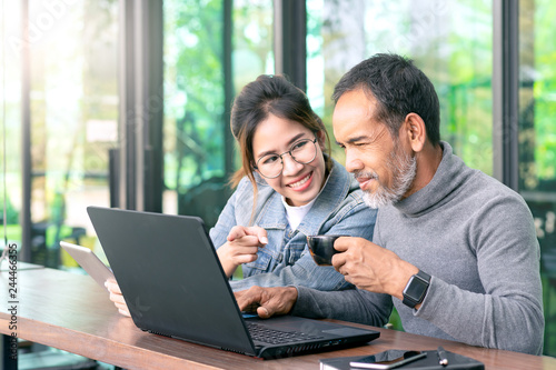 Attractive mature asian man with white stylish short beard looking at laptop computer with teenage eye glasses hipster woman in cafe. Teaching internet online or wifi technology in older man concept.