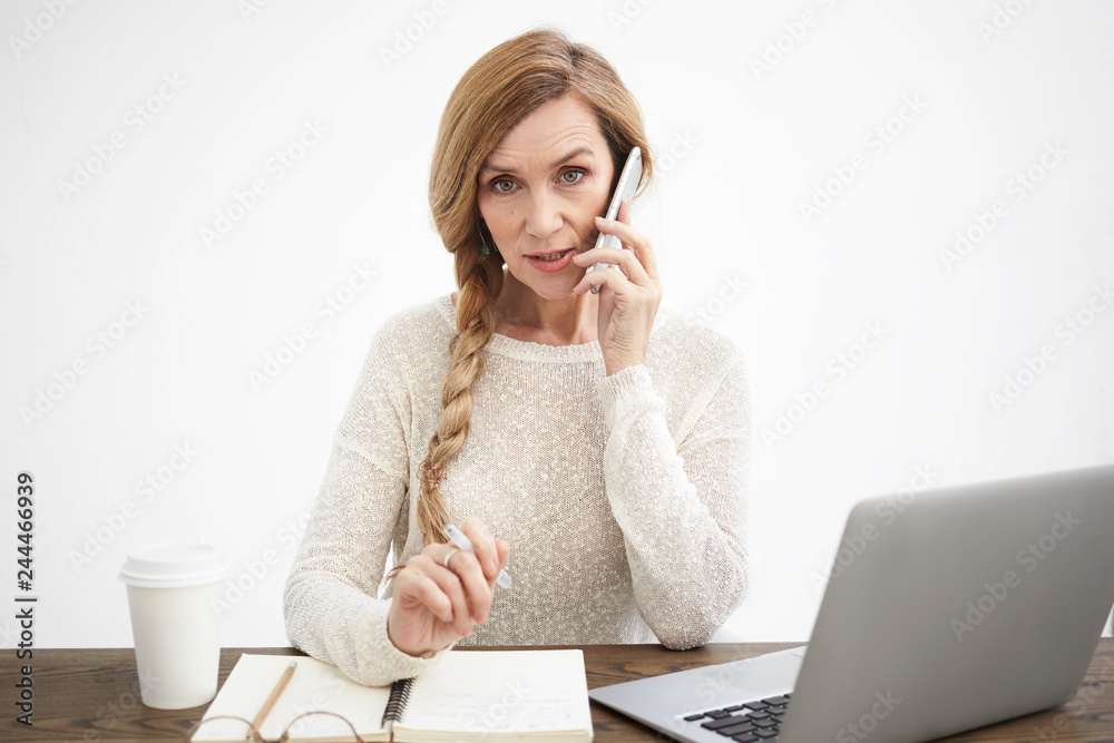 Beautiful elderly businesswoman with natural make up and long colored hair gathered in braid speaking on smart phone while working at desk, holding pen, writing down important notes in copybook Beautiful elderly businesswoman with natural make up and long colored hair gathered in braid speaking on smart phone while working at desk, holding pen, writing down important notes in copybook