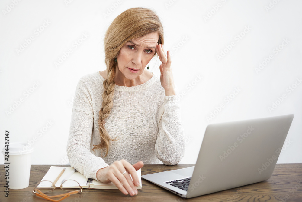 People, age, job, occupation and technology concept. Portrait of tired mature female copywriter with braid having painful look because of stress or deadline, sitting in front of open laptop at desk People, age, job, occupation and technology concept. Portrait of tired mature female copywriter with braid having painful look because of stress or deadline, sitting in front of open laptop at desk