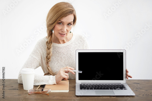 Cheerful stylish mature sixty year old female presenting new product or concept, looking at camera and pointing pen at laptop screen with copyspace for your information or advertising content