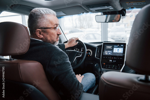 Front devices are on. View from behind of senior businessman in official clothes driving a modern new car