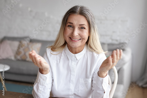 Human facile expressions, positive emotions and feelings. Horizontal shot of beautiful happy retired woman with loose dyed blonde hair with gray roots, gesturing emotionally and smiling at camera