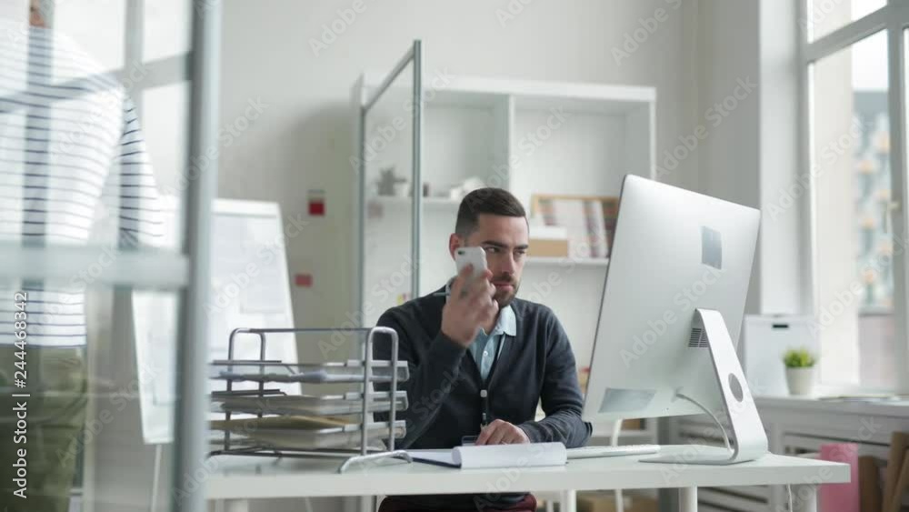 Tracking right of businessman with beard sitting at desk, writing and then answering telephone call, while his colleague passing with papers in his hands