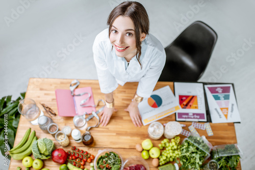 Portrait of a young woman nutritionist in medical uniform standing near the table full of various healthy products indoors photo
