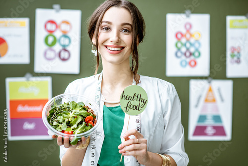 Portrait of a woman dietitian in medical gown standing with salad on the green wall background with drawings on a topic of healthy food indoors