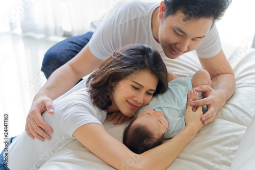 Asian father and mother are hugging new newborn baby on sofa. They are smiling and warm touching to the baby with love. Father holding both of baby hand and mother shoulder. Happy family concept.