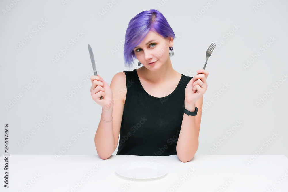 Fork, plate, knife - Concept portrait of a beautiful girl with purple hair on a white background sitting at a table with dining tools. Fork, plate, knife - Concept portrait of a beautiful girl with purple hair on a white background sitting at a table with dining tools.