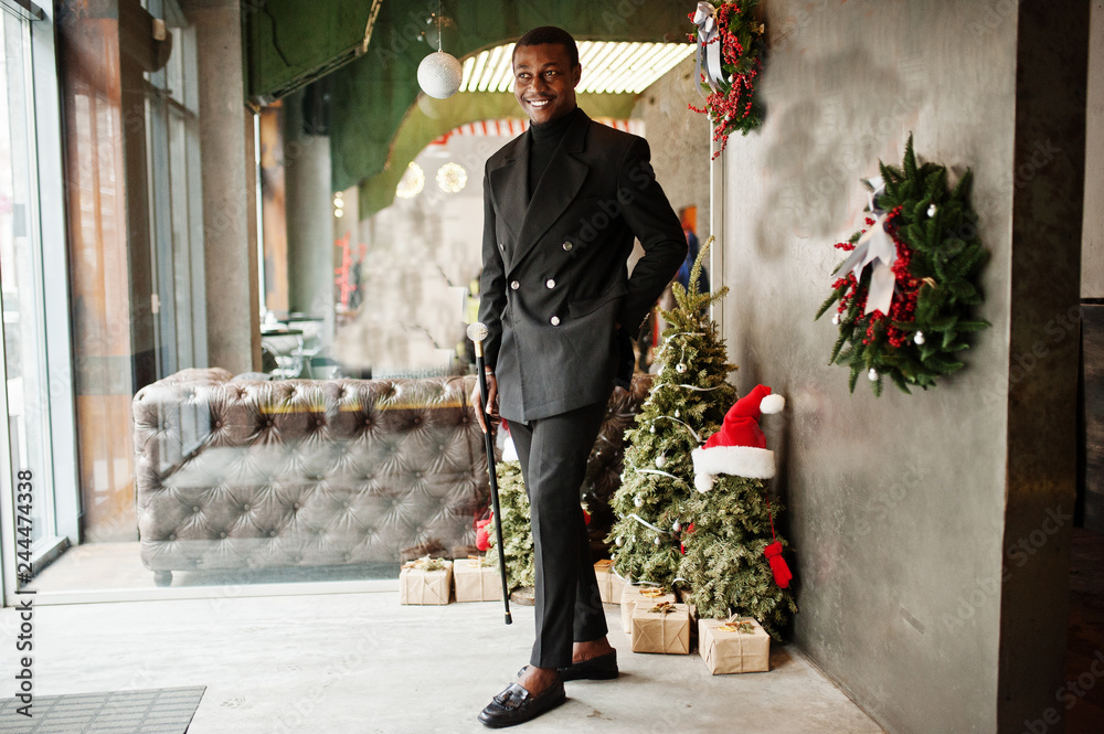 african american gentleman in elegant black jacket, holding retro walking  stick as cane flask or tippling cane with golden diamond ball handle. Rich  fashionable afro man against Christmas decorations. Stock Photo