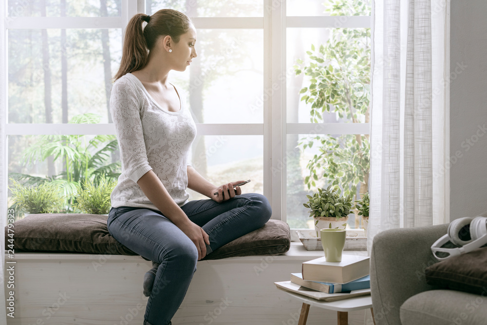 Young woman sitting on the windowsill and using her phone Young woman sitting on the windowsill and using her phone