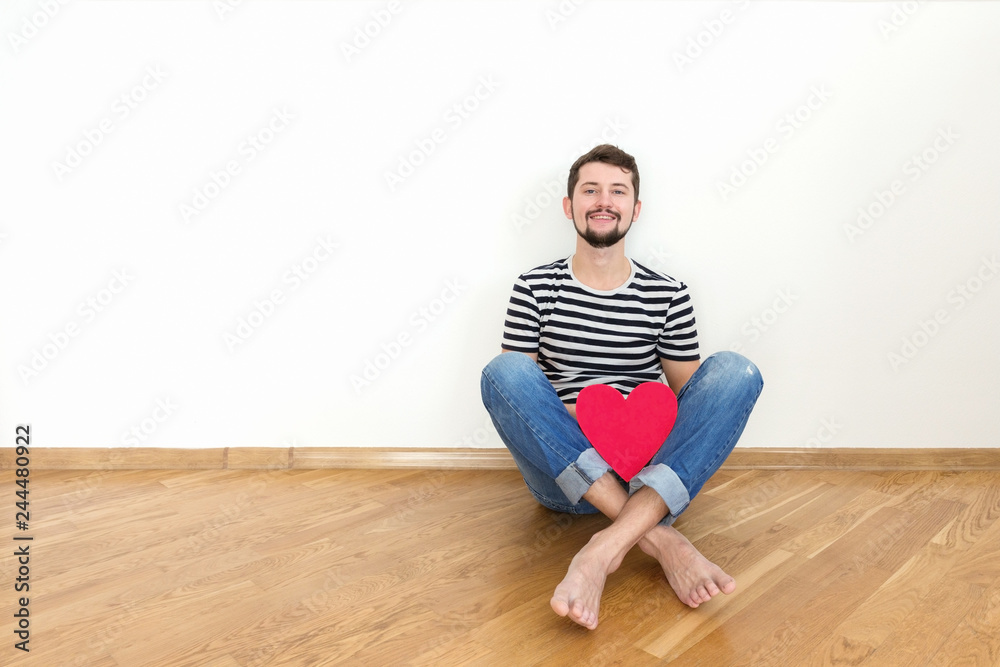 Happy valentines day! Young man is sitting on the floor with red heart and smiling. Copy space. Happy valentines day! Young man is sitting on the floor with red heart and smiling. Copy space.