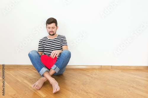 Happy valentines day! Young man is sitting on the floor with red heart and smiling. Copy space.