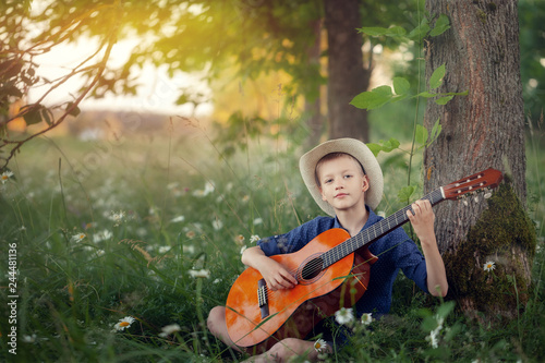 Adorable boy with guitar, relaxing in the park. Kid sitting on a grass in summer day