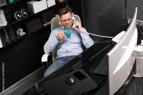 man leaned back in his chair, drinking coffee and talking on landline photo