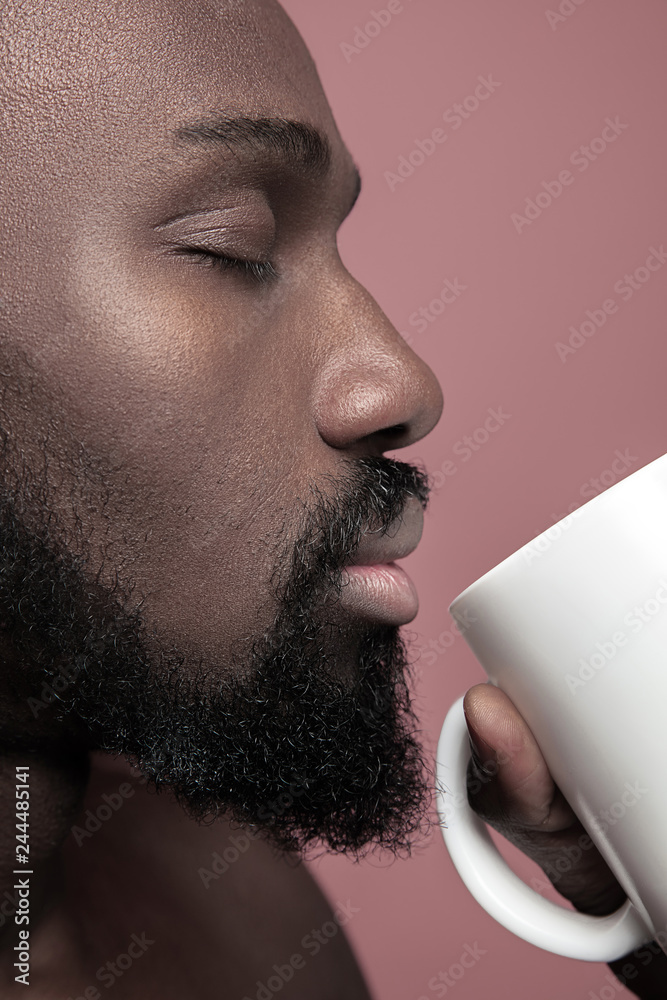 African man with white cup of tea, isolated on pink studio background. Close up portrait in minimalism style of a young naked happy afro man - obrazy, fototapety, plakaty 