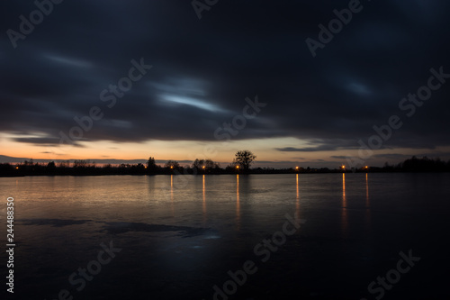 Cloudy dark clouds after sunset on a frozen lake and reflecting lights on the ice
