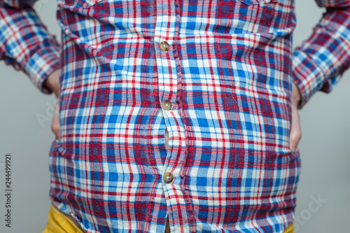 fat man in a plaid shirt is having trouble fastening his small clothes with buttons; his bare belly is visible through the holes