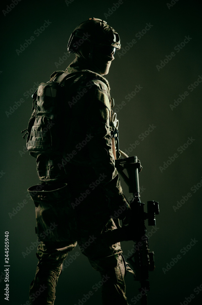 Low key studio shoot of army special forces soldier, commando fighter in mask, ballistic glasses, tactical helmet and battle uniform, holding short barrel service rifle, looking back over shoulder Low key studio shoot of army special forces soldier, commando fighter in mask, ballistic glasses, tactical helmet and battle uniform, holding short barrel service rifle, looking back over shoulder