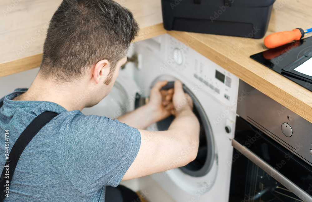 Professional handyman in overalls repairing washing machine in the kitchen. Professional handyman in overalls repairing washing machine in the kitchen.