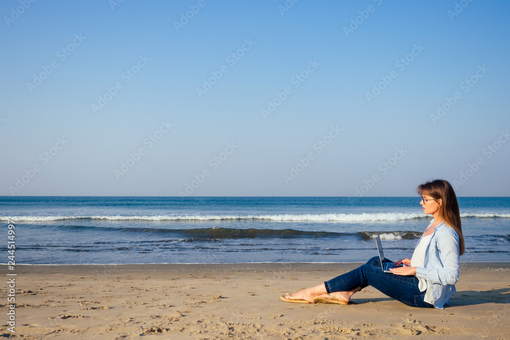 Young sexy woman in stylish glasses using laptop freelancing on the tropical paradise beach. Girl freelancer work sitting in a summer sand on the seashore remote work indian ocean copyspace success