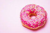 Bright donut in a pink glaze with a multi-colored rainbow sprinkle on a pink background. 