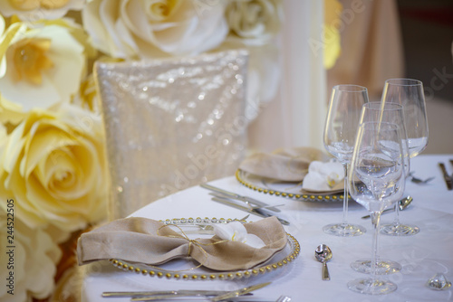 Sitting arrangement at a formal event or fine dining restaurant featuring transparent plates with golden details, glassware and silverware in the order of use set against a paper flowers wall © Ana
