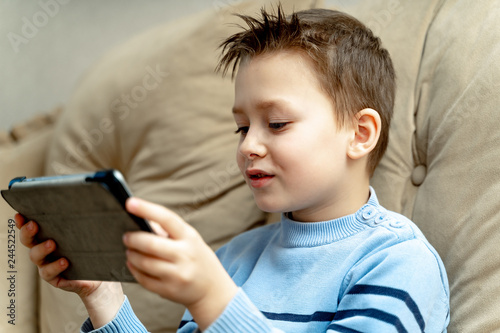 Happy boy of school age in blue sweater sitting on sofa with wireless tablet. Kid relaxing at home playing games on tablet after back from school. Close-up