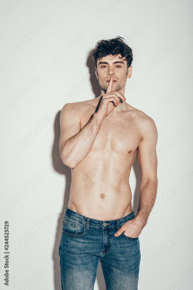 sexy shirtless man in jeans showing silence symbol while posing on grey