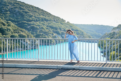 The beautiful girl in a blue dress and sunglasses poses on the bridge, the long chestnut hair, happy and smiles, azure water of the lake and slopes of mountains on a background © Vladimir Drozdin