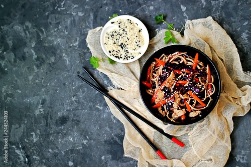 Wheat noodles stir fry with red cabbage, carrots and turkey in a black bowl. Served with black and white sesame seeds. Chinese food. Top view, copy space.