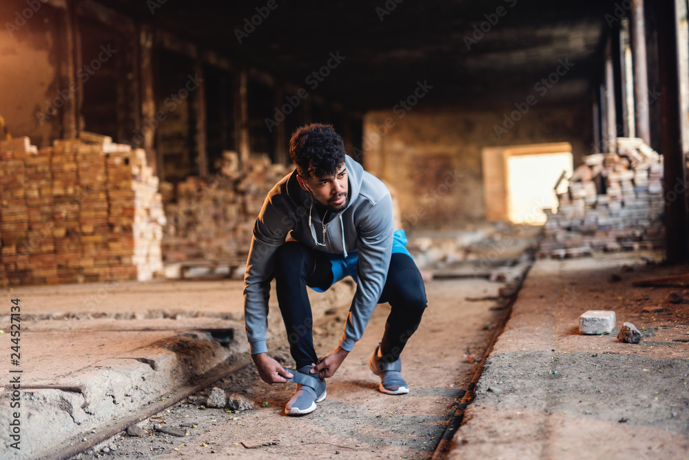 Portrait of african american guy putting on sneaker while crouching in old brick factory. Headphones on. Portrait of african american guy putting on sneaker while crouching in old brick factory. Headphones on.