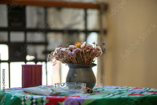 Vase of beautiful dried flowers standing on table with colorful tablecloth next to cigarettes, ash tray and lighter. Concept of unhealthy morning habits. 