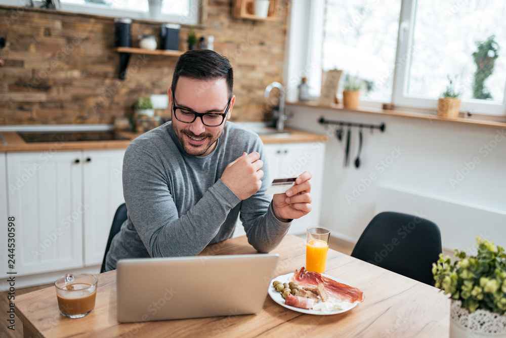 Young man sitting at the kitchen table with breakfast in morning using laptop and credit card. Young man sitting at the kitchen table with breakfast in morning using laptop and credit card.