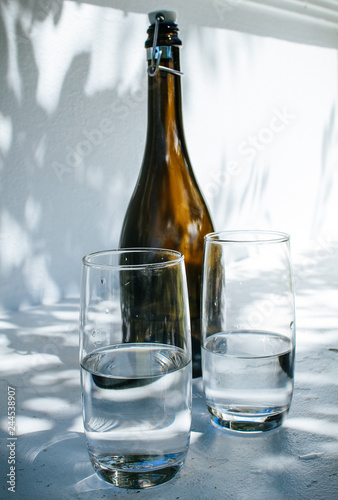 One bottle and two glasses of water standing on a white rim against a bright white wall with shadows of leaves on a sunny summer day