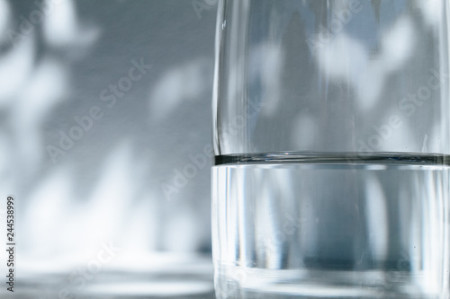 Close up of a glass of water standing in front of a white wall with shadows of leaves on a bright, sunny day