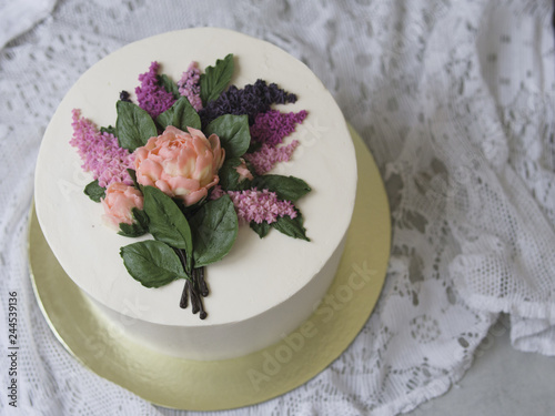 White cream cake decorated with buttercream flowers - lilac, peonies - on wooden background with lace fabric. Vintage, retro. Gift for Women's Day, 8 March, Vaentine's Day. Close up, copy space