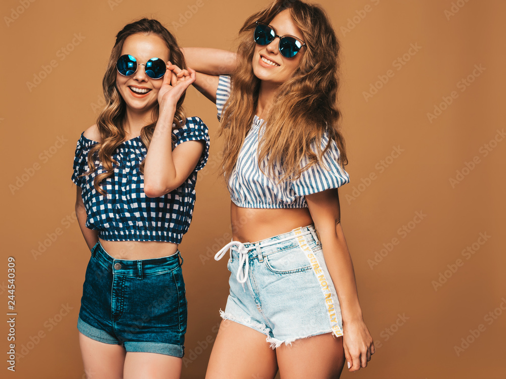 Two young beautiful smiling hipster girls in trendy summer clothes and sunglasses. Sexy carefree women posing on golden background in studio. Positive models Two young beautiful smiling hipster girls in trendy summer clothes and sunglasses. Sexy carefree women posing on golden background in studio. Positive models