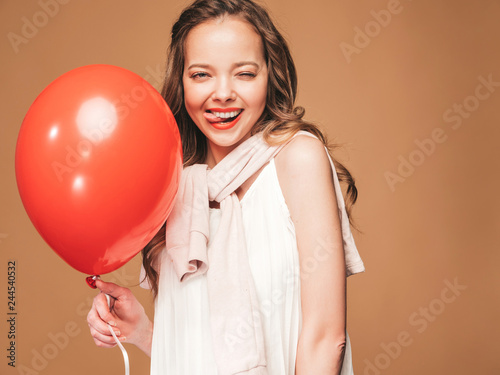 Excited young girl posing in trendy summer white dress. Woman model with red air balloon posing on golden background.Showing her tongue and ready for party © halayalex