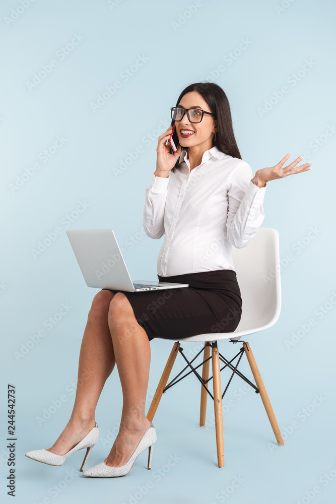 Pregnant business woman isolated over blue wall background using laptop computer talking by mobile phone. Pregnant business woman isolated over blue wall background using laptop computer talking by mobile phone.