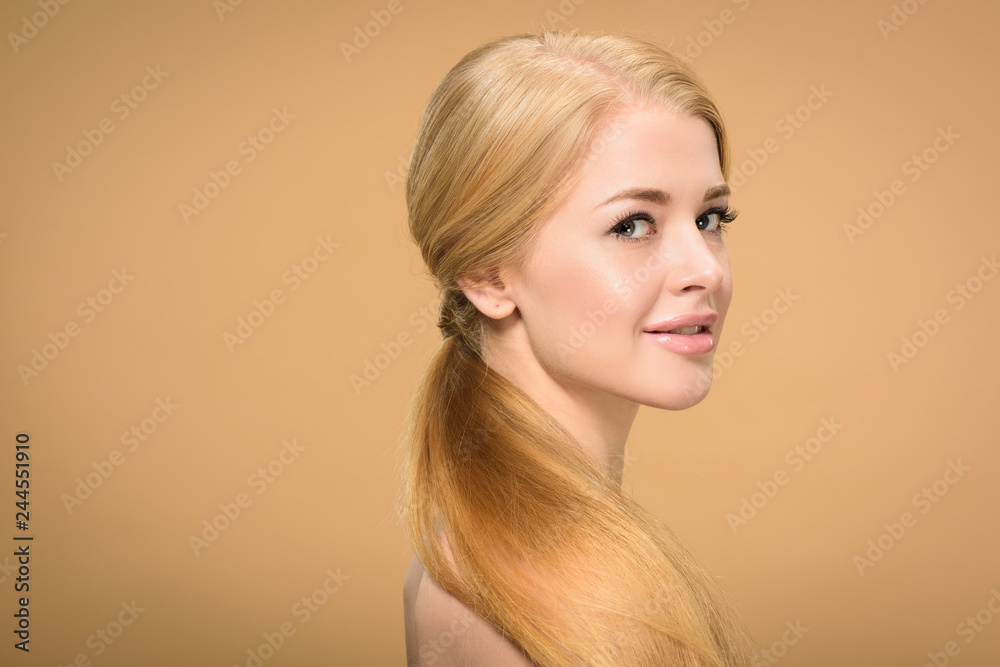 Fototapeta premium side view of beautiful young blonde woman with long hair smiling at camera isolated on beige