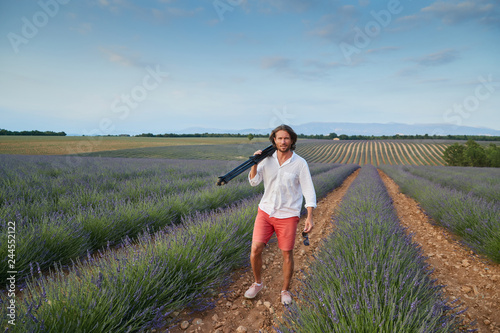 The beautiful young brutal man goes to a lavender field at sunset, he is dressed in a white shirt with a short sleeve and red shorts, the photographer holds a tripod, he smiles © Vladimir Drozdin