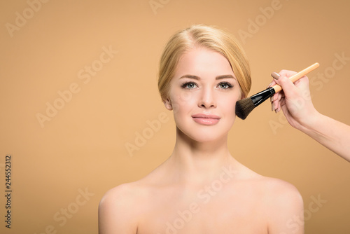 cropped shot of stylist applying makeup with cosmetic brush to young naked woman looking at camera isolated on beige
